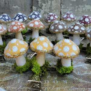May include: A collection of decorative mushroom figurines with white polka dots on the caps. The caps are various colors, including orange, yellow, purple, and brown, with gold glitter trim. The mushrooms are set on a bed of green moss.