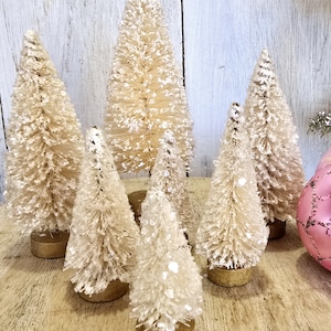 May include: A set of seven white bottle brush Christmas trees with gold bases. The trees are arranged in a row on a wooden surface.