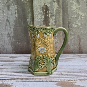 May include: A ceramic pitcher featuring a hexagonal body with a mustard yellow background and raised relief patterns of green leaves and white flowers. The piece includes a curved green handle and a molded spout, showcasing a detailed botanical design.