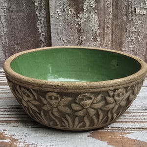 May include: A round ceramic bowl with a smooth, vibrant green interior glaze and a textured, unglazed tan exterior. The outer surface features a repeating embossed floral pattern of daffodils encircling the entire circumference of the bowl.