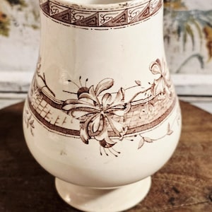 May include: Cream-colored ceramic vase with brown floral and geometric patterns. The vase has a bulbous body, a pedestal base, and a slightly flared rim. The floral design features stylized flowers and leaves.