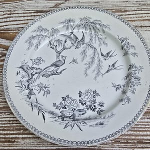 May include: A round ceramic plate featuring a black and white transferware pattern of birds perched on branches, flying birds, and blooming flowers. The rim is decorated with a repeating circular border design on a cream-colored background.