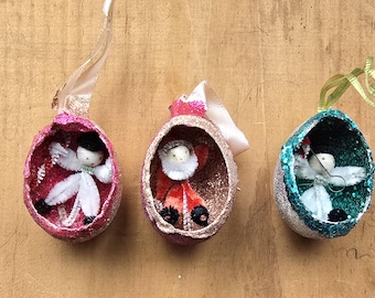 May include: Three vintage Christmas ornaments. Each ornament is egg-shaped, with a glittery exterior in pink, gold, and teal. Inside each egg is a small figure, including an angel and Santa Claus. Each ornament has a ribbon for hanging.
