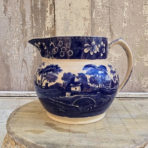 May include: A blue and cream-colored ceramic pitcher with a landscape design. The pitcher features a detailed blue transferware pattern of trees, buildings, and a bridge. The rim is decorated with floral motifs.
