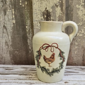 May include: A cream-colored ceramic pitcher featuring a central illustration of a brown chicken standing inside a green pine wreath tied with a red ribbon. The pitcher has a rounded body, a short spout, and a curved handle on the right side.