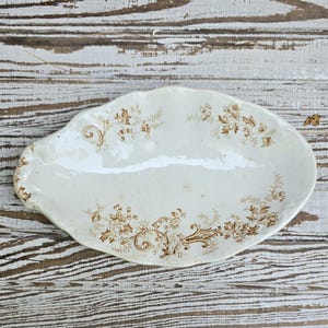 May include: An oval-shaped ceramic dish with a scalloped edge, featuring a cream-colored base and a brown floral transferware pattern. The design includes delicate botanical motifs and scrolling vines distributed across the surface of the tray.