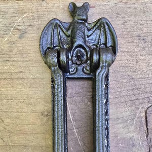 May include: A black cast iron door knocker in the shape of a bat with outstretched wings. The bat is centered above a rectangular frame with vertical bars. The door knocker is mounted on a wooden surface.
