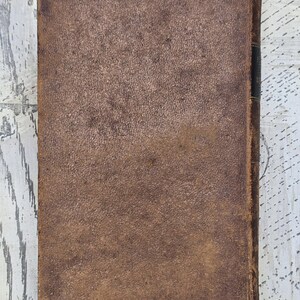 Puede incluir: Un libro antiguo con una cubierta de cuero marr&oacute;n desgastado. La cubierta tiene una apariencia texturizada con desgaste visible, lo que sugiere edad y uso. El lomo es oscuro y los bordes muestran signos de edad.