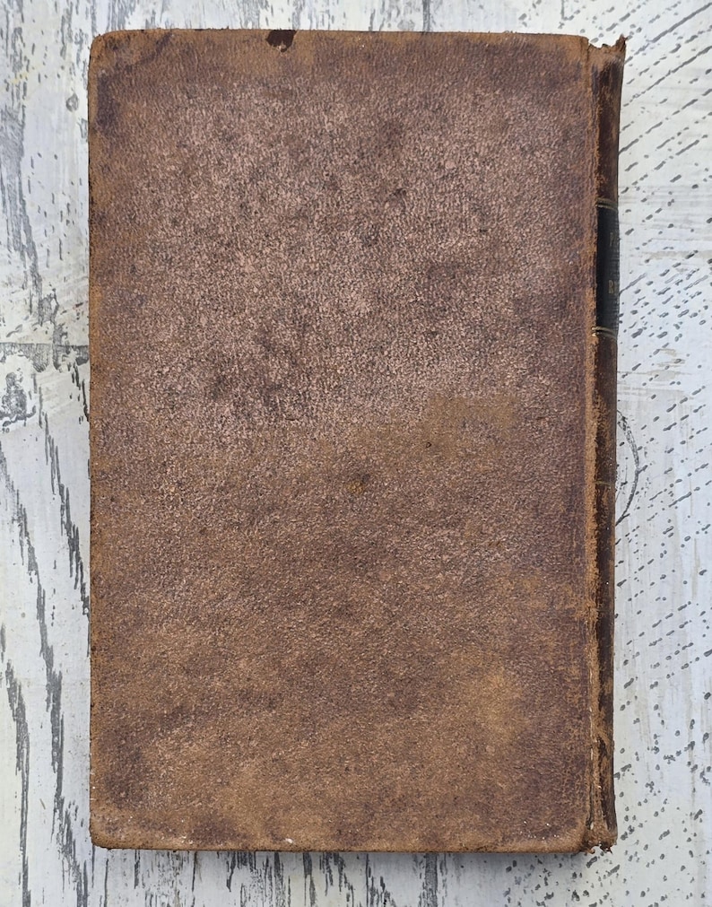 Puede incluir: Un libro antiguo con una cubierta de cuero marr&oacute;n desgastado. La cubierta tiene una apariencia texturizada con desgaste visible, lo que sugiere edad y uso. El lomo es oscuro y los bordes muestran signos de edad.