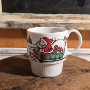 May include: White ceramic Christmas mug featuring Santa Claus in a red suit and hat, riding a sleigh with gifts. The mug has a handle and a decorative base. The design includes a green Christmas tree and candy canes.