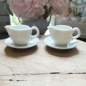 May include: Two small, white ceramic cream pitchers with handles and matching saucers. The pitchers have a classic design with a spout for easy pouring. The set is displayed on a light-colored wooden surface, with a blurred background of pink flowers.