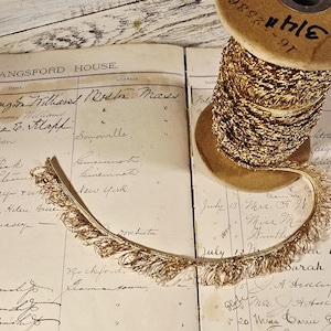 May include: A spool of gold-colored chain and a length of matching fringe sit on a vintage ledger. The ledger's pages are filled with handwritten entries. The spool is marked with the number "16-2536".