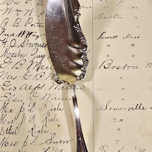 May include: A vintage silver cake server with an ornate handle and a wide, curved blade. The server is set against a backdrop of aged paper with handwritten text, suggesting a historical or antique theme.
