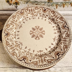 May include: A vintage ceramic plate with a cream-colored base and intricate brown floral patterns. The plate features a central floral medallion and a decorative border. This antique plate is a decorative piece for the home.