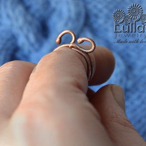 Op de afbeelding: Een ring van koperdraad met een uniek ontwerp. De ring wordt aan een vinger gedragen en heeft twee lussen die met elkaar zijn gedraaid.