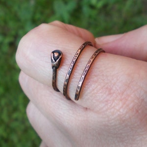 May include: A copper ring with a textured finish and a small, black, flower-shaped accent. The ring is wrapped around a finger.