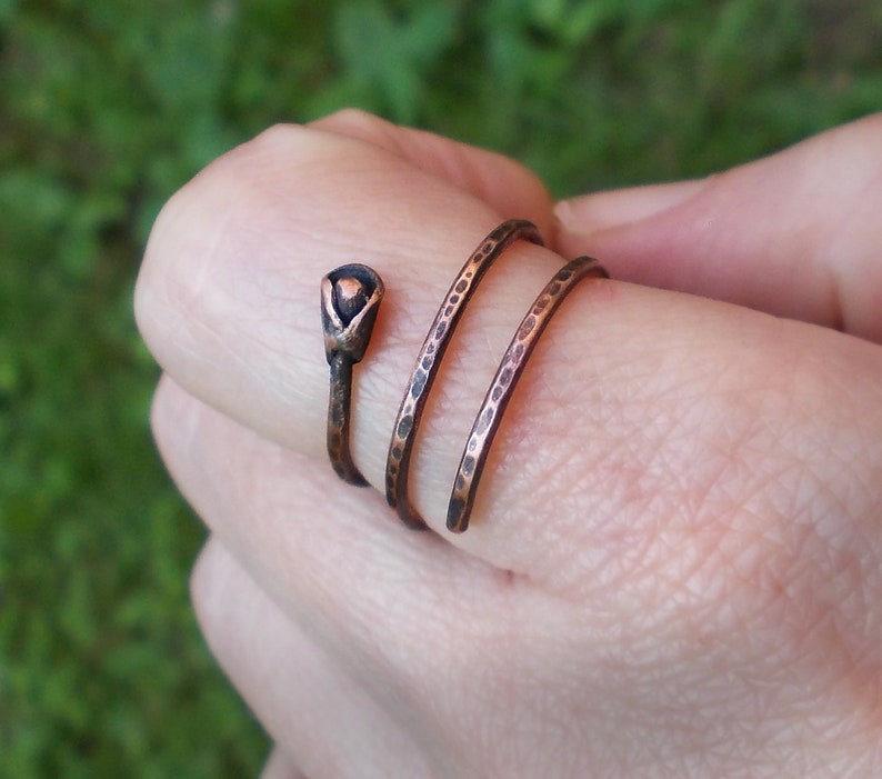May include: A copper ring with a textured finish and a small, black, flower-shaped accent. The ring is wrapped around a finger.