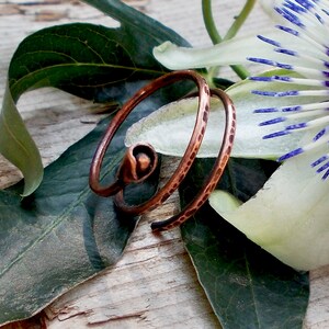May include: A copper wire ring with a textured finish. The ring is coiled and has a small bead at the end. The ring is photographed on a wooden surface with green leaves and a white flower.