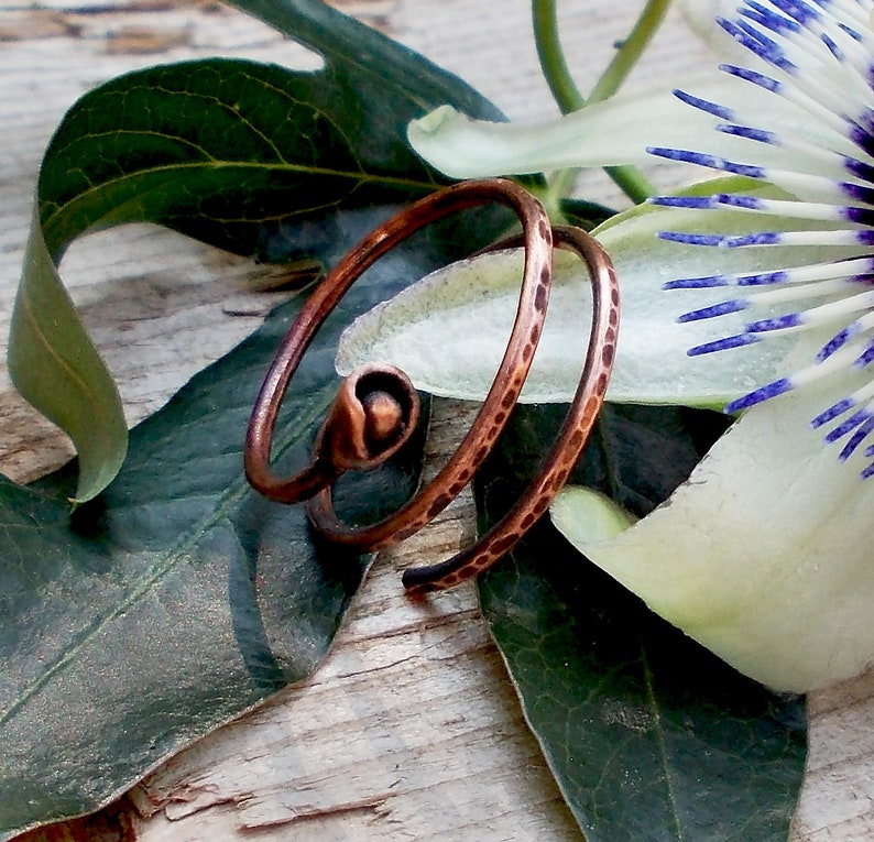 May include: A copper wire ring with a textured finish. The ring is coiled and has a small bead at the end. The ring is photographed on a wooden surface with green leaves and a white flower.