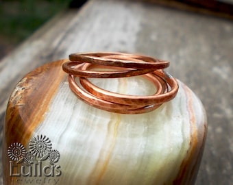 May include: A set of four hammered copper rings stacked on a light brown and white stone surface. The rings are a warm copper colour and have a textured, hammered finish.