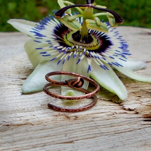 May include: A copper wire ring with a twisted design and a small flower accent. The ring is lying on a wooden surface next to a white flower with blue and purple accents.