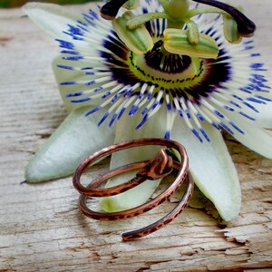 May include: A copper wire ring with a spiral design. The ring is lying on a wooden surface next to a white flower with blue and purple accents.