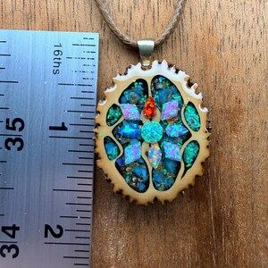 May include: A handcrafted pendant necklace featuring a mosaic design. The pendant, approximately 1.5 inches in diameter, showcases a vibrant arrangement of blue, green, and iridescent stones set within a light brown, textured frame. The necklace has a tan cord.
