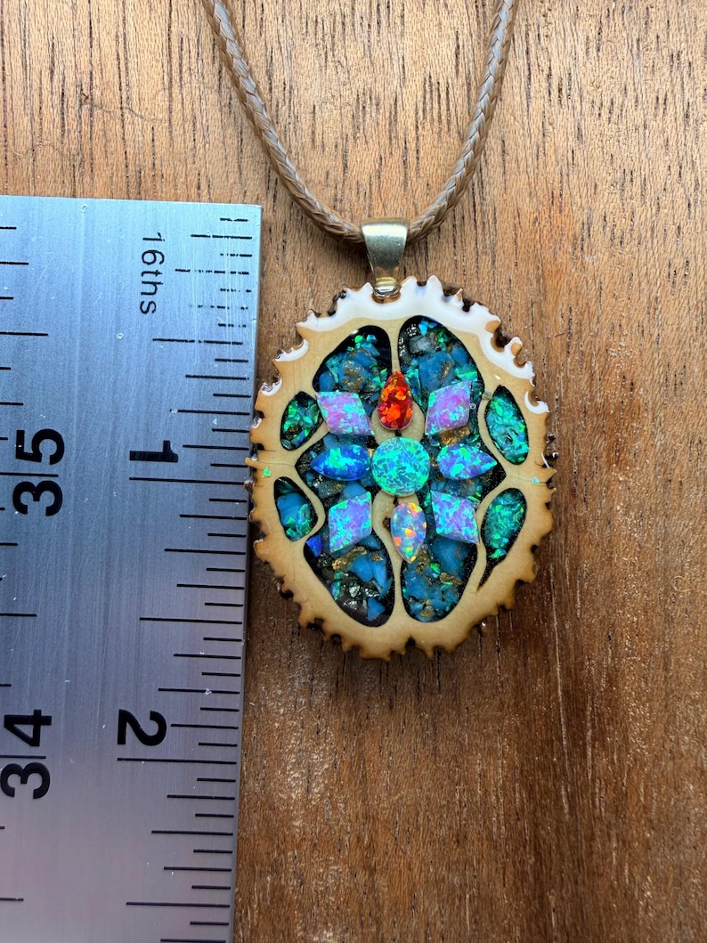 May include: A handcrafted pendant necklace featuring a mosaic design. The pendant, approximately 1.5 inches in diameter, showcases a vibrant arrangement of blue, green, and iridescent stones set within a light brown, textured frame. The necklace has a tan cord.