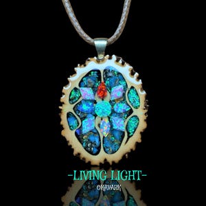 May include: A handcrafted pendant necklace featuring a mosaic design with blue, green, and red gemstones set in a light brown, circular frame. The pendant hangs from a light brown cord. The text "-LIVING LIGHT- OKAVARK" is displayed below the pendant.