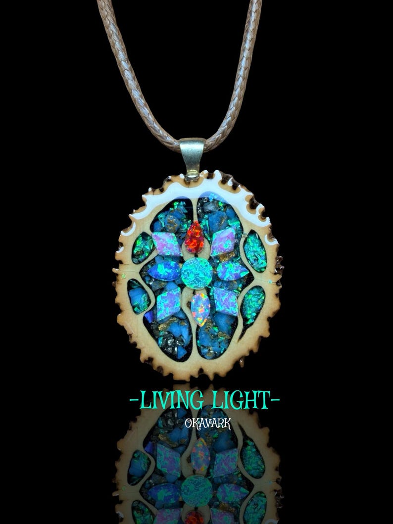 May include: A handcrafted pendant necklace featuring a mosaic design with blue, green, and red gemstones set in a light brown, circular frame. The pendant hangs from a light brown cord. The text "-LIVING LIGHT- OKAVARK" is displayed below the pendant.