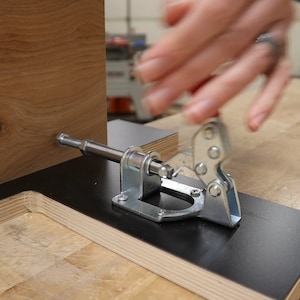 以下が含まれることがあります： 黒い表面に付けられた銀色のクランプのクローズアップ。クランプは木片を固定するために使用されています。木は薄い茶色で、節があります。