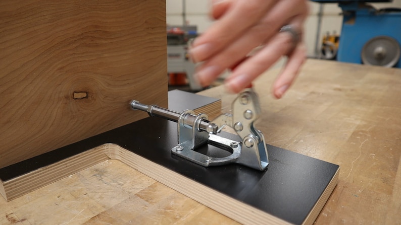 以下が含まれることがあります： 黒い表面に付けられた銀色のクランプのクローズアップ。クランプは木片を固定するために使用されています。木は薄い茶色で、節があります。