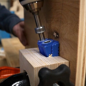 以下が含まれることがあります： 青いポケットホールジグが木材に穴を開けるために使用されている様子のクローズアップ。ジグは木材にクランプされ、ドリルを使用してポケットホールを作成しています。