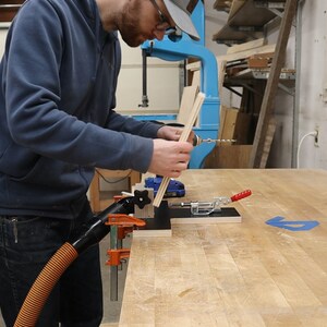 以下が含まれることがあります： 人がドリルを使って木片を別の木片に固定しています。木片はクランプで作業台に固定されています。ドリルには集塵機が接続されており、木くずを吸い込んでいます。