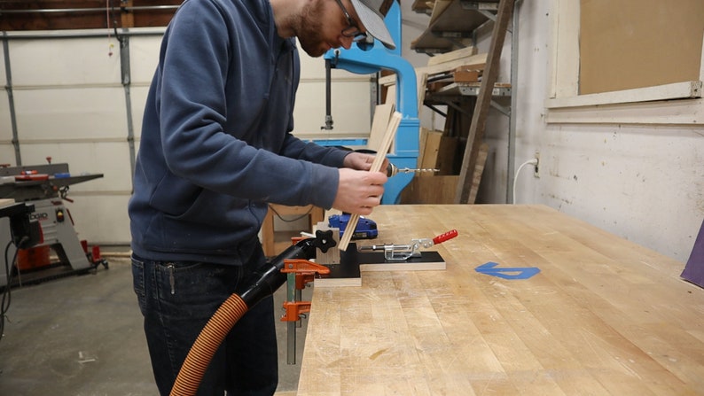 以下が含まれることがあります： 人がドリルを使って木片を別の木片に固定しています。木片はクランプで作業台に固定されています。ドリルには集塵機が接続されており、木くずを吸い込んでいます。