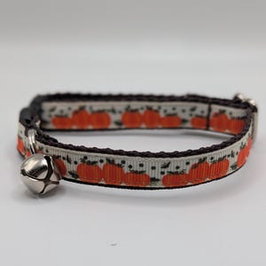 May include: A pet collar featuring a white ribbon with a repeating pattern of orange pumpkins and small black dots. The collar is edged in dark brown fabric and includes a silver-toned metal bell attached to a metal ring on the left side.
