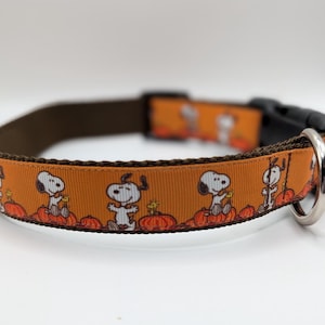 May include: An orange pet collar featuring a repeating pattern of Snoopy and Woodstock sitting on pumpkins. The collar has brown fabric edges, a black plastic side-release buckle, and a silver-toned metal D-ring for attaching a leash or tags.