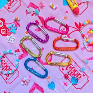 Puede incluir: Una colección de mosquetones de colores rosa, morado, naranja, dorado y azul, con el texto "+ Jemma +" en cada uno. La imagen también incluye pulseras de cuentas, pendientes con forma de caramelo y una tela rosa con la frase "GOOD VIBES".