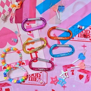 Puede incluir: Un conjunto de cinco mosquetones ovalados de metal en morado, naranja, dorado, azul y rosa, grabados con la palabra QUEER y estrellas. Incluye dos pulseras de cuentas, cuatro pendientes con forma de piruleta y pequeños dijes de estrellas.
