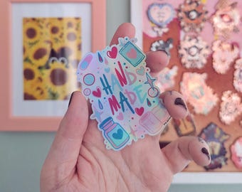 Può includere: Un adesivo olografico con le parole "HAND MADE" in lettere colorate. L'adesivo presenta varie illustrazioni, tra cui cuori, pillole e un flacone con un cuore. L'adesivo è tenuto in una mano.