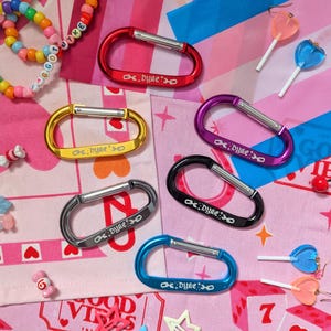 May include: Six metal carabiners in red, gold, purple, black, silver, and blue, each featuring the word "dyke" in white text. The collection includes a multi-coloured beaded bracelet, four heart-shaped lollipop earrings, and three star-shaped hair clips.