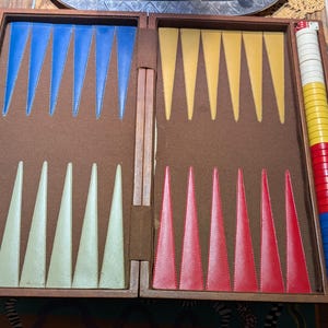 May include: A wooden backgammon board with a brown felt interior featuring blue, cream, yellow, and red triangular points. A stack of white, yellow, red, and blue checkers sits on the side next to a ruler measuring approximately 18 inches in length.