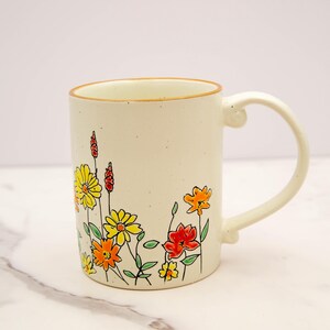 May include: Cream-colored ceramic mug with a floral design. The mug features a curved handle and a rim with a gold-colored trim. The floral design includes yellow, orange, and red flowers with green stems and leaves. The mug is approximately 4 inches tall.