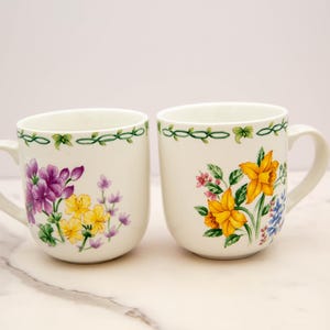 May include: Two white ceramic mugs with floral designs. One mug features purple and yellow flowers, while the other has yellow and blue flowers.