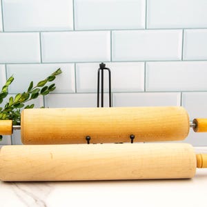 May include: Two wooden rolling pins with wooden handles are displayed on a black metal stand against a white tiled background.