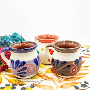 May include: Three handcrafted ceramic mugs with unique designs. One mug has a white base with blue and purple floral patterns, another has a white base with brown floral patterns, and the third has a white base with yellow polka dots. Each mug has a brown rim and handle.