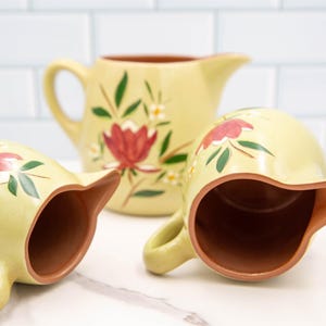 Puede incluir: Tres jarras de cerámica amarilla con diseños florales. Las jarras tienen un interior marrón y están decoradas con flores rojas y verdes.