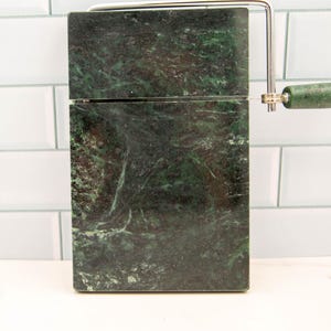 Könnte beinhalten: Ein grünes Marmor-Käsebrett mit einem eingebauten Draht-Käseschneider. Das Brett hat ein dunkelgrünes und weißes Marmormuster.