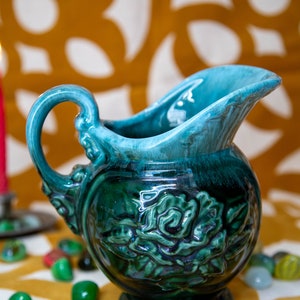 Könnte beinhalten: Eine grüne und blaue Keramikkanne mit Blumenmuster. Die Kanne hat einen Henkel und eine breite Öffnung.