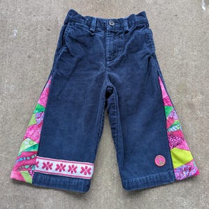 May include: Navy blue corduroy pants featuring a button closure, belt loops, and side pockets. The outer leg panels are decorated with a colorful patchwork of pink, green, and purple fabric. A white band with pink flowers and a peace sign patch.